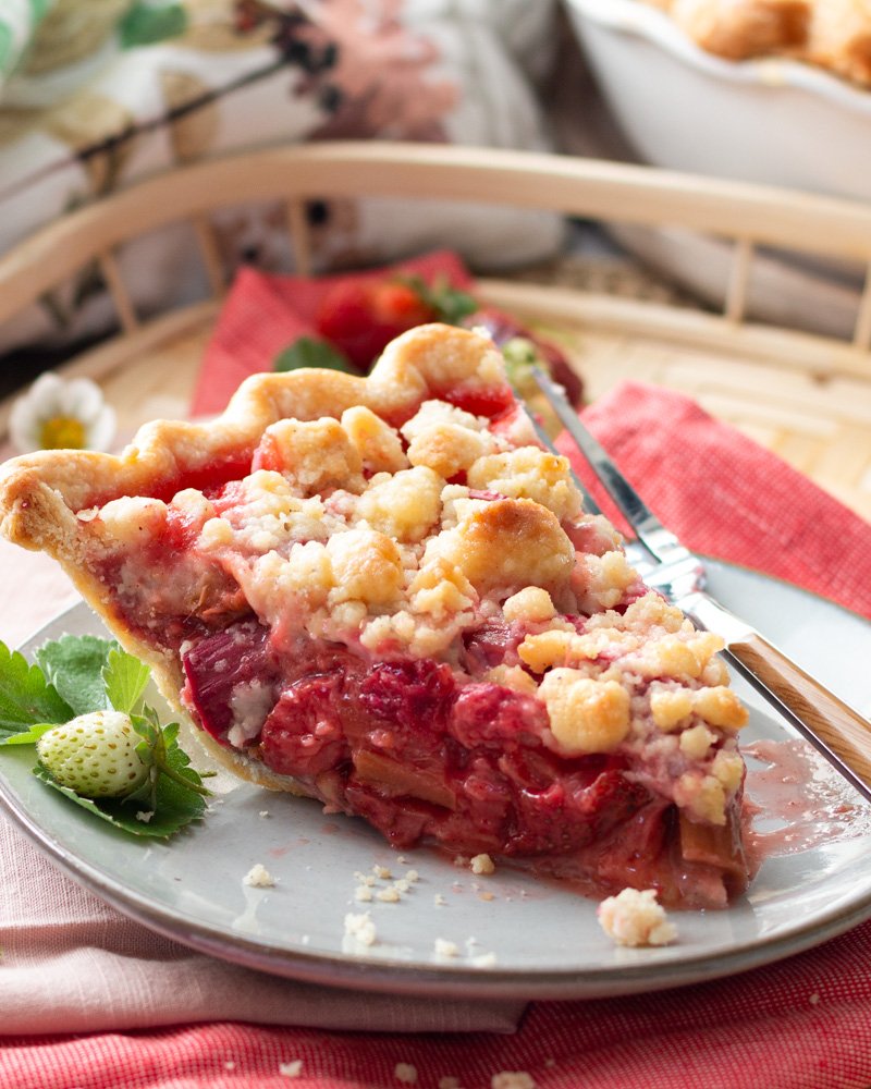 A strawberry rhubarb pie with a slice cut out on a basket and strawberry leaves and flowers.