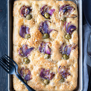 Tuscan focaccia in a baking sheet that's golden brown with sliced red onion and green olive.